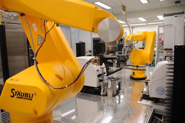 A robot arm (foreground) retrieves assay plates from incubators and places them at compound transfer stations or hands them off to another arm (background) that services liquid dispensers or plate readers.